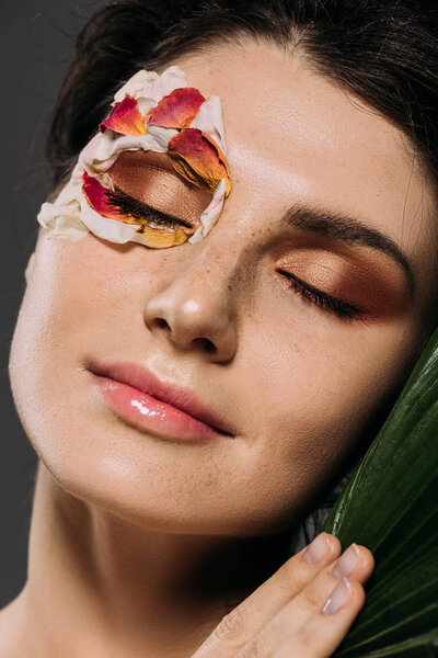 beautiful young woman with closed eyes with makeup and floral petals on face holding leaf isolated on grey