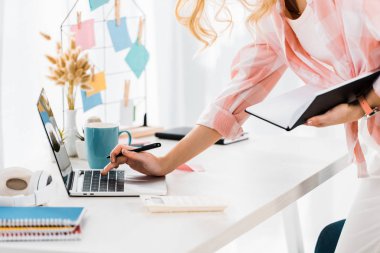 Partial view of woman with notebook and pen typing on laptop keyboard clipart