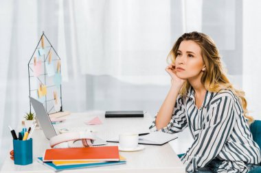Pensive young woman in striped shirt sitting at workplace and propped face with hand clipart