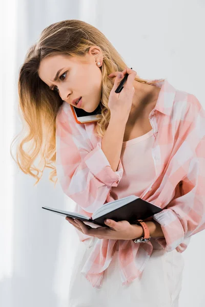Confused Young Woman Holding Notebook Talking Smartphone — Stock Photo, Image