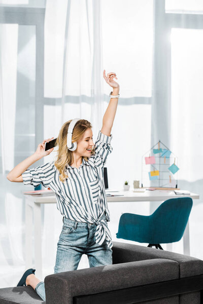 Blissful girl in striped shirt with smartphone dancing and listening music in headphones