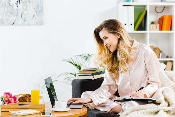 Lachende Vrouw Pyjama Schrijven Notitieboekje Laptop Thuis Gebruiken — Stockfoto