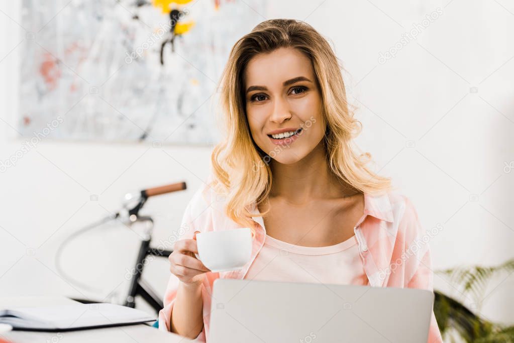 Beautiful young woman with laptop drinking coffee and looking at camera