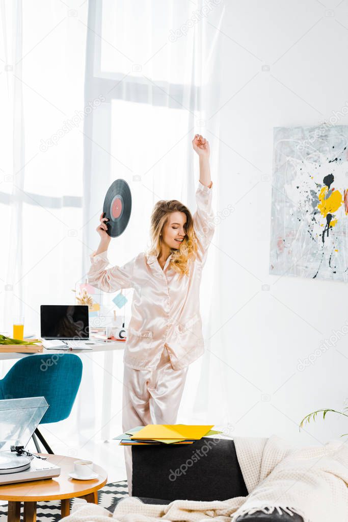 Happy girl in pyjamas holding vinyl record and dancing in morning