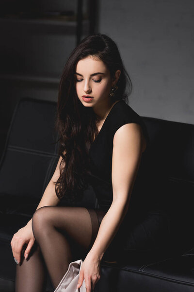 attractive brunette woman in elegant dress sitting on couch on dark background