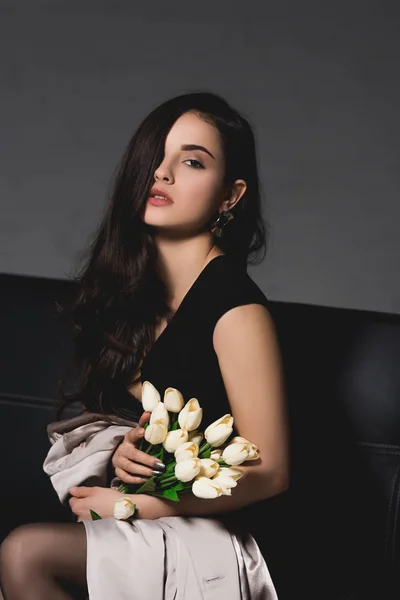 Привлекательная Брюнетка Черном Платье Сидит Диване Держит Белые Тюльпаны Темном — стоковое фото
