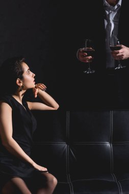 Kanepenin duran ve gözlük şarap ile siyah arka plan üzerine tutarak siyah elbiseli Wile yakışıklı adam oturan esmer kadın