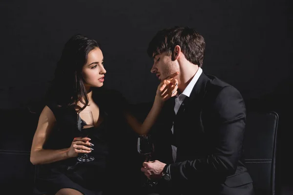 黒のドレスと黒い背景にガラスとソファに座っているスーツでハンサムな男の魅力的なブルネットの女性 — ストック写真