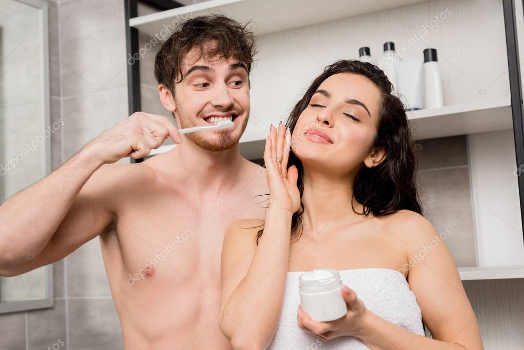 Attractive woman holding cream and man brushing teeth in bathroom