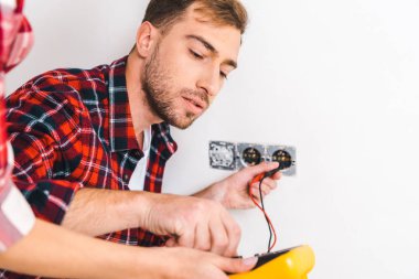 cropped view of woman holding digital multimeter near handsome boyfriend sitting near power socket and holding cables clipart