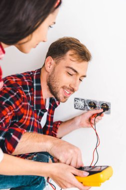selective focus of man looking at digital multimeter while holding cables near girlfriend at home clipart