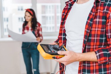 selective focus of man holding digital multimeter with woman holding blueprint on background  clipart