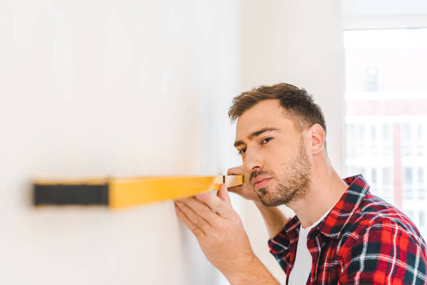 handsome handyman holding measuring level while measuring wall at home