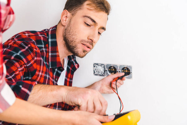 cropped view of woman holding digital multimeter near handsome boyfriend sitting near power socket and holding cables