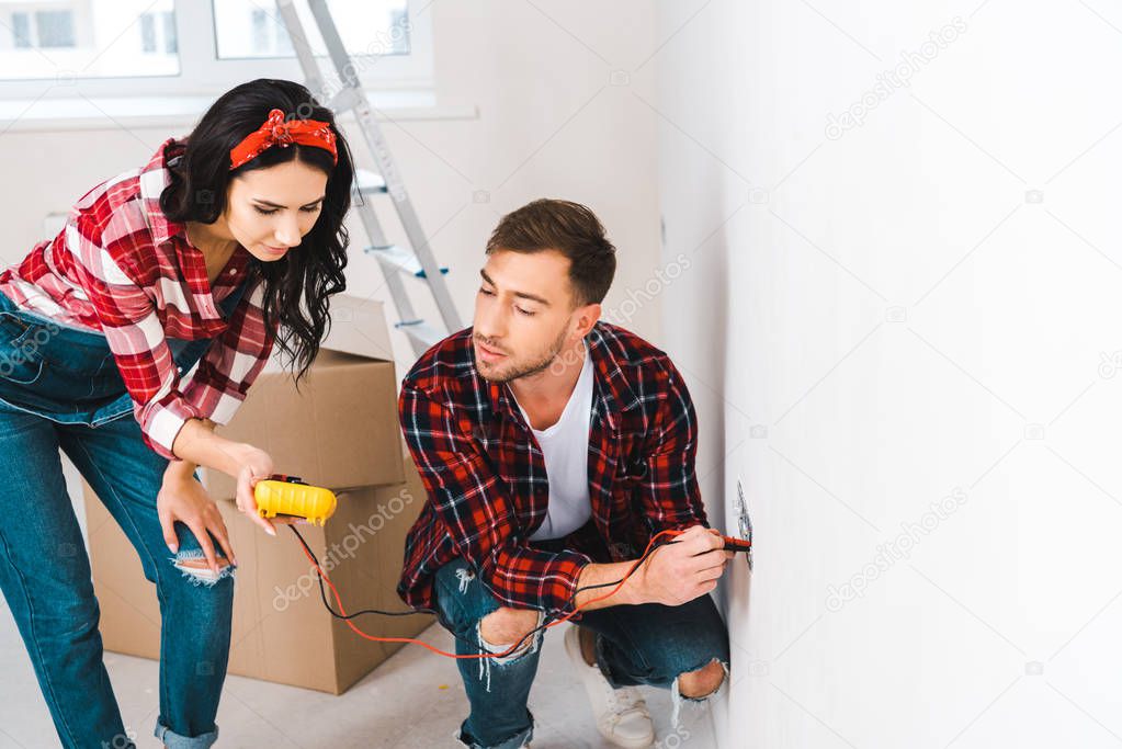 attractive woman holding digital multimeter near boyfriend sitting near power socket and holding cables