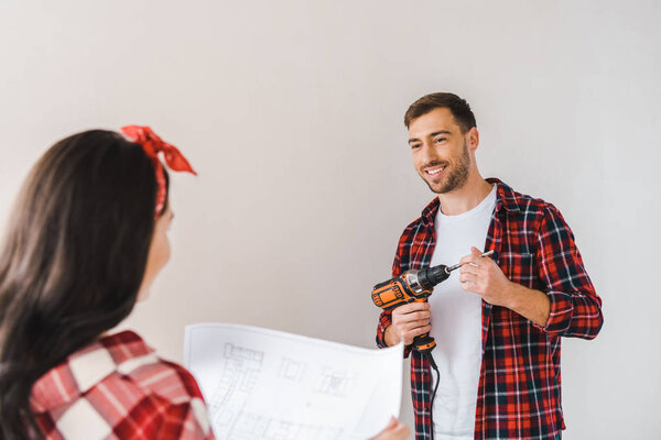 happy man holding drilling machine and looking at woman holding architecture plan on paper in hands