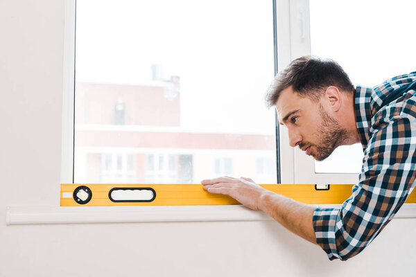 handsome handyman holding measuring level near window in room