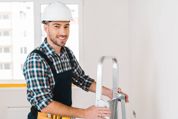 cheerful handyman in helmet smiling while holding ladder 