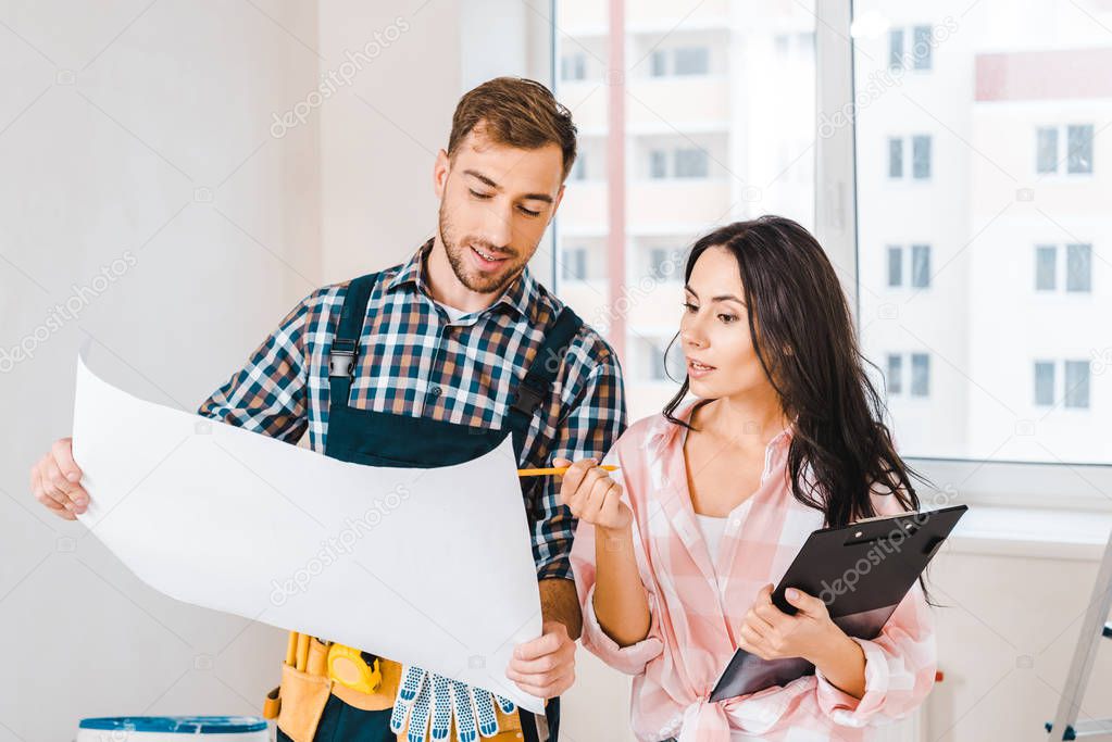 cheerful handyman holding blueprint near attractive client with clipboard and pencil