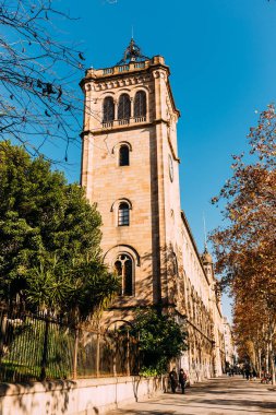 BARCELONA, SPAIN - DECEMBER 28, 2018: street with green trees and high old tower on blue sky background clipart