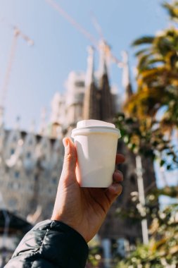 BARCELONA, SPAIN - DECEMBER 28, 2018: selective focus of male hand with white paper cup, blurred Temple Expiatori de la Sagrada Familia on background clipart