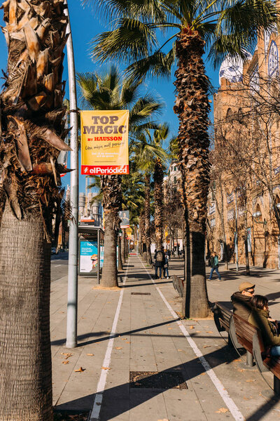 BARCELONA, SPAIN - DECEMBER 28, 2018: city street with tall green palm trees and people sitting on benches