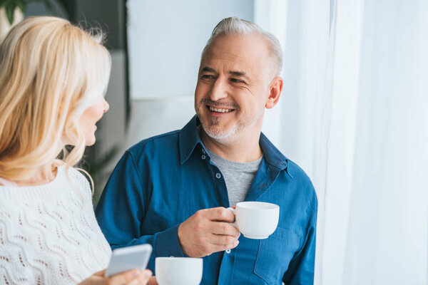 cheerful man holding cup and smiling while looking at wife with smartphone