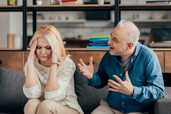 selective focus of woman holding head while sitting on sofa near emotional husband 