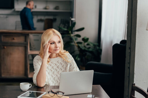 избирательный фокус усталой женщины, держащей голову, сидя рядом с ноутбуком с мужем на заднем плане
