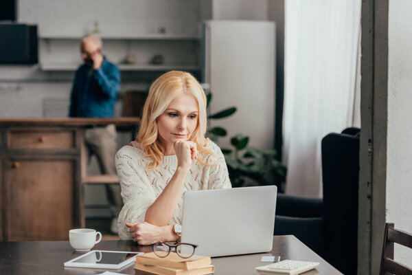 избирательный фокус задумчивой женщины, сидящей рядом с ноутбуком с мужем на заднем плане
