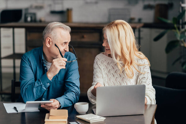 красивый мужчина держит очки и цифровой планшет, глядя на привлекательную жену с ноутбуком
