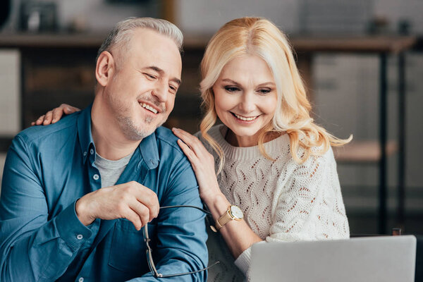 handsome man holding glasses and smiling near attractive wife while looking at laptop