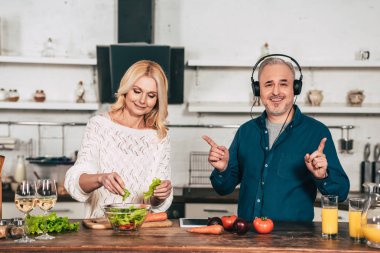cheerful woman preparing food near happy husband listening music in headphones in kitchen clipart