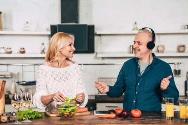 happy woman preparing food near happy husband listening music in headphones in kitchen clipart