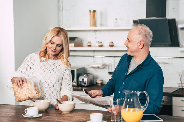 happy man holding smartphone and newspaper near wife making breakfast in kitchen