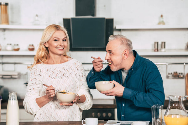 attractive wife holding bowl with cornflakes near cheerful husband eating in kitchen