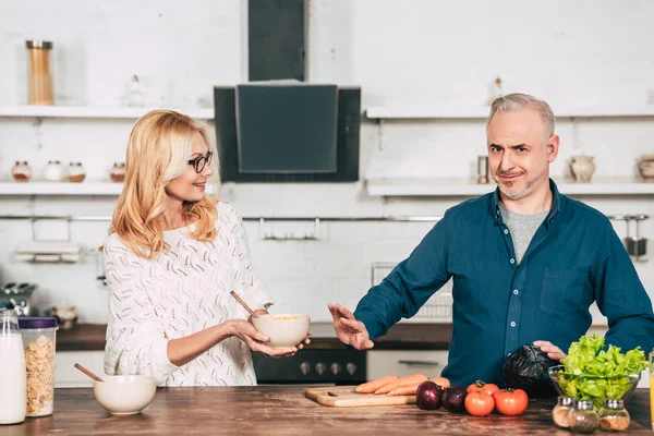 Atractiva Mujer Gafas Sosteniendo Tazón Cerca Marido Gesto Cocina —  Fotos de Stock