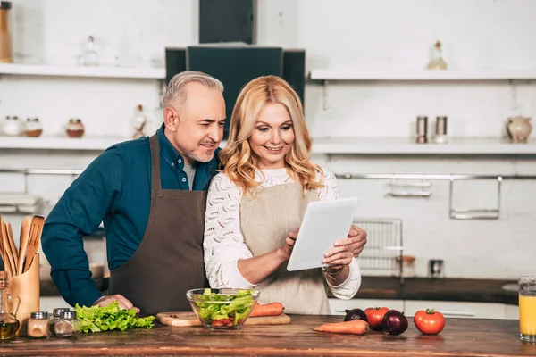 Szczęśliwa Kobieta Pomocą Cyfrowego Tabletu Stojąc Mężem Kuchni Pobliżu Warzyw — Zdjęcie stockowe