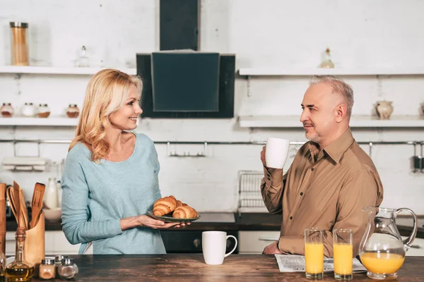 happy wife holding plate with croissants near husband with cup of drink