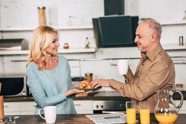 cheerful wife holding plate with croissants near husband with cup of drink
