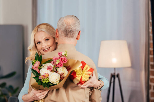 счастливая женщина улыбается, обнимая мужа и держа в руках цветы и подарочную коробку
