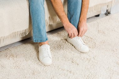 adam oturan ve evde spor ayakkabılar üzerinde ayakkabı bağcığı bağlama Jeans görünümünü kırpılmış