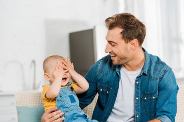 elleriyle gözlerini kapsayan ve gülümseyen baba evde CE oyun oynuyor yürümeye başlayan çocuk