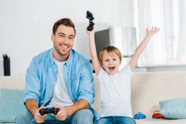 preschooler son cheering with hands in air while playing video game with father at home clipart