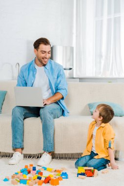 kanepede oturan ve renkli yapı taşları ile evde oynarken preschooler oğlu ise dizüstü kullanarak gülümseyen baba