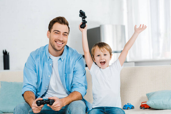 preschooler son cheering with hands in air while playing video game with father at home