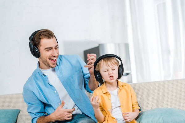 father with preschooler son in headphones listening music, gesturing with hands and playing imaginary guitars at home 