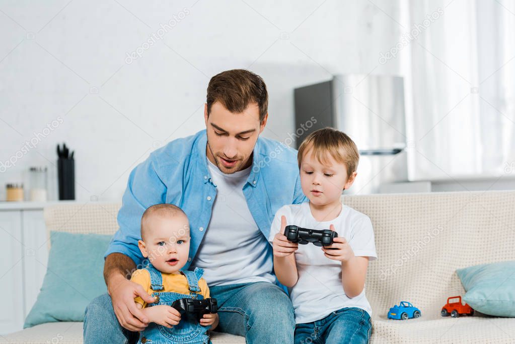 father playing video game with preschooler and toddler sons while sitting on couch at home