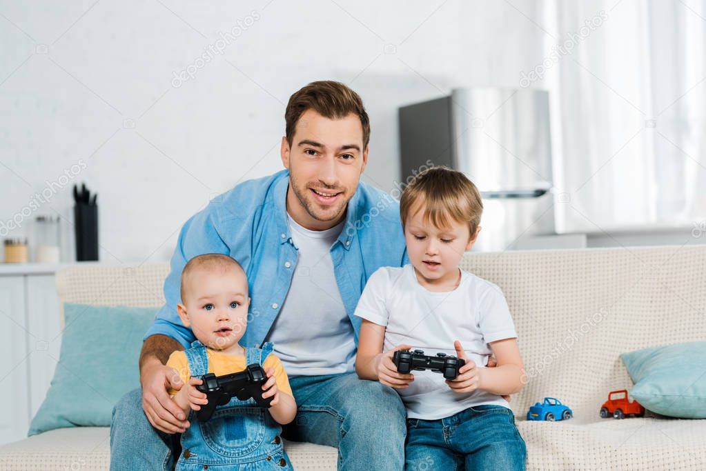 smiling father playing video game with adorable preschooler and toddler sons while sitting on couch at home