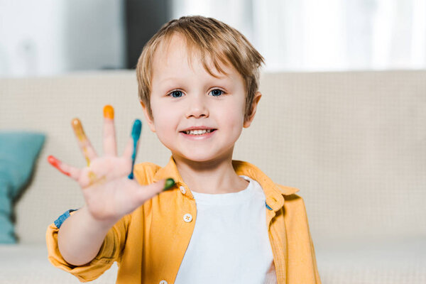 adorable smiling preschooler boy with colorful paint on hand looking at camera at home